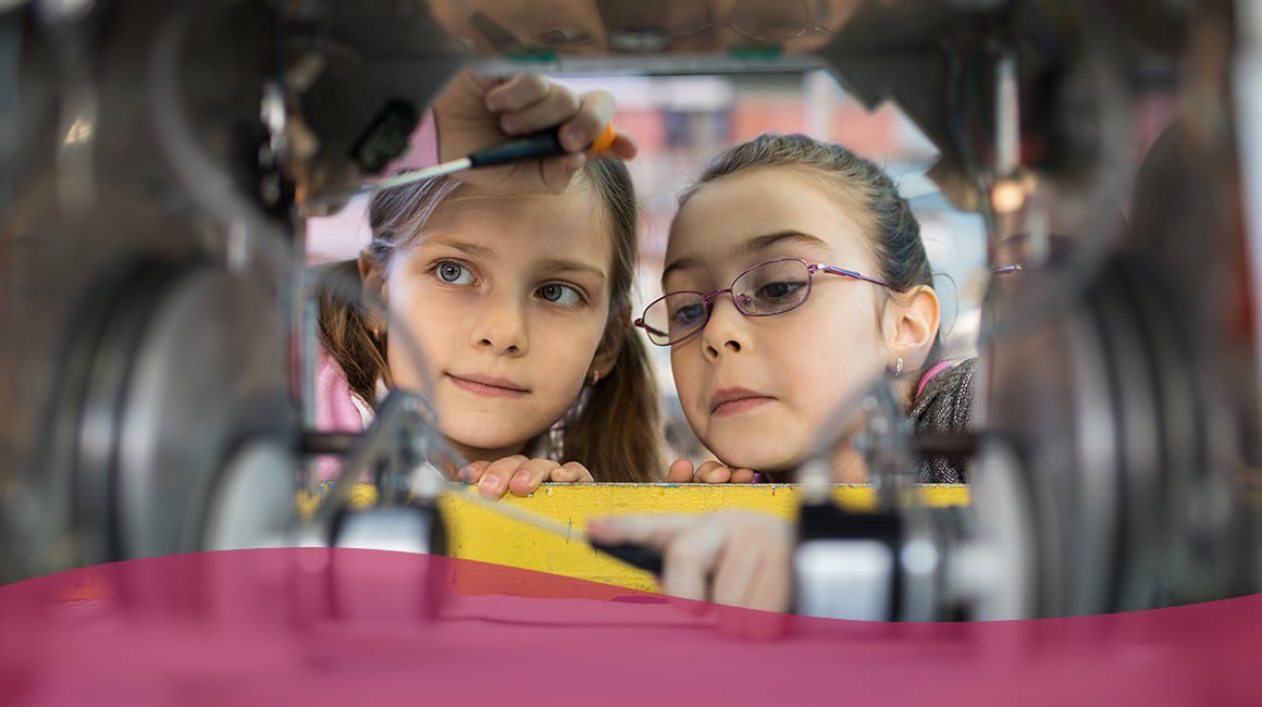 Two young girls observing the inside of a mechanical device.