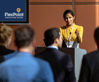 A woman behind a podium in front of an audience. She is giving a presentation to them.
