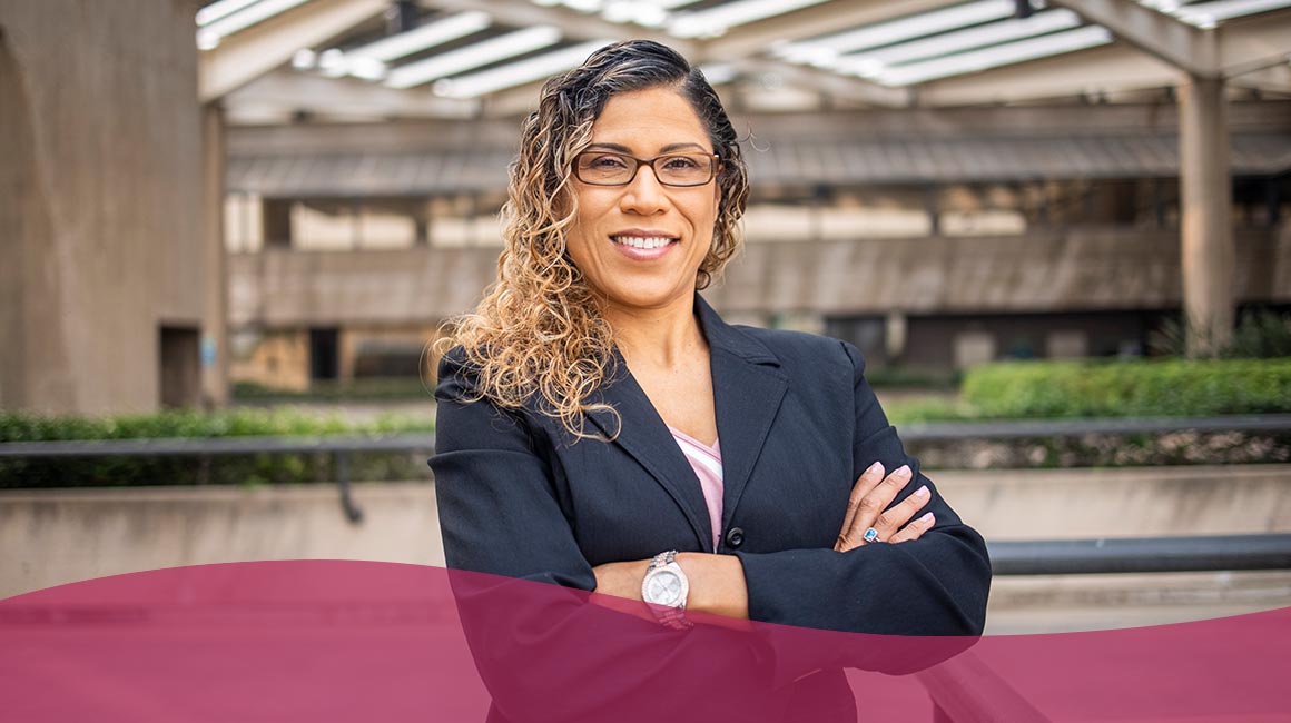 A person in a dark blazer with glasses stands confidently outside with architectural background.