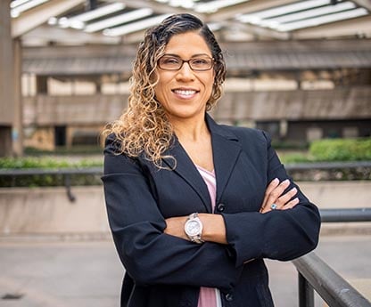 A person in a dark blazer with glasses stands confidently outside with architectural background.