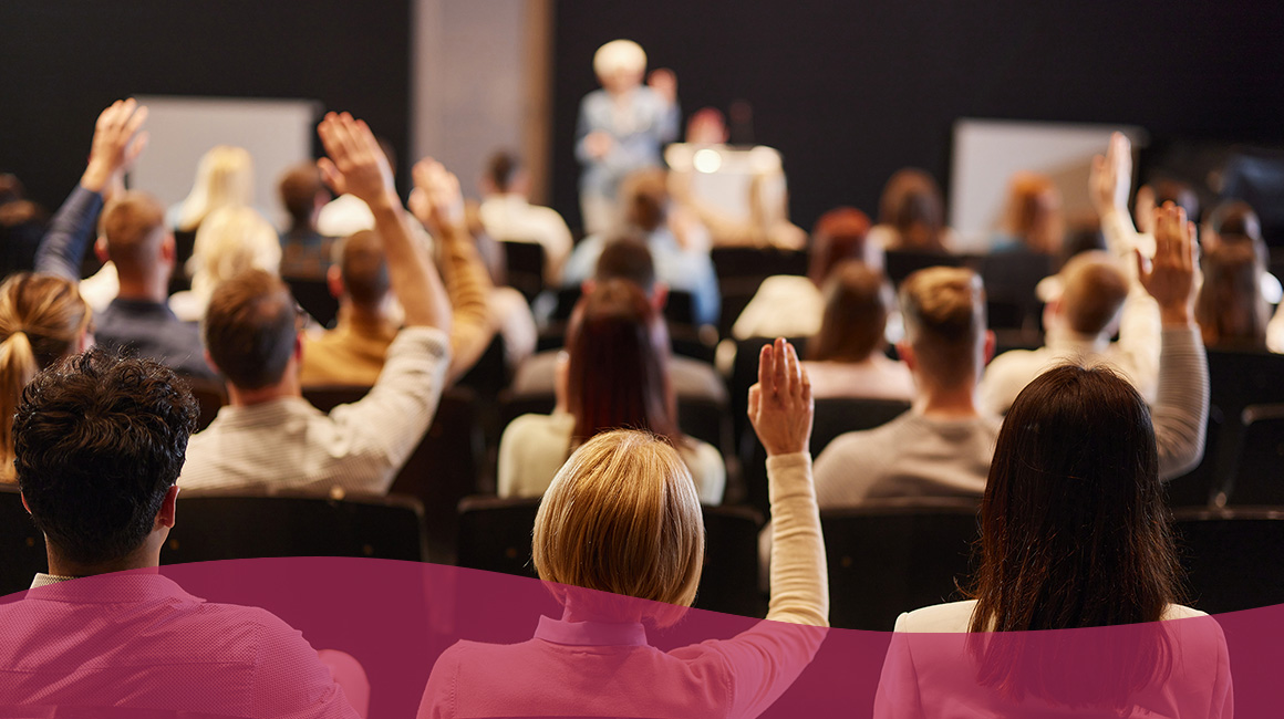 A group of people at a conference all raising their hands while staring at a stage where someone is talking