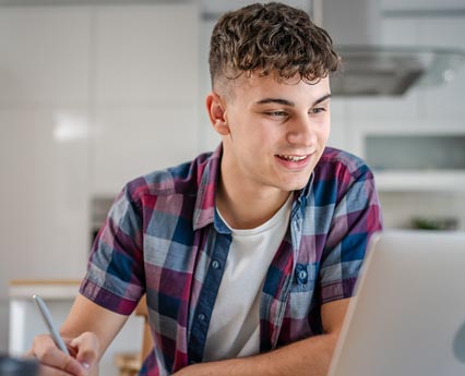 A young person attending a virtual school smiling at a laptop, wearing a blue and pink plaid shirt in a kitchen setting.