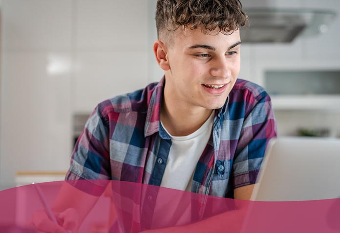 A young person attending a virtual school smiling at a laptop, wearing a blue and pink plaid shirt in a kitchen setting.