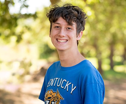 Thrive School student, Waldo, in a park surrounded by trees wearing a blue shirt and smiling