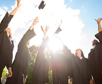 A group of graduating students stand in a circle with the graduation robes, tossing their caps.